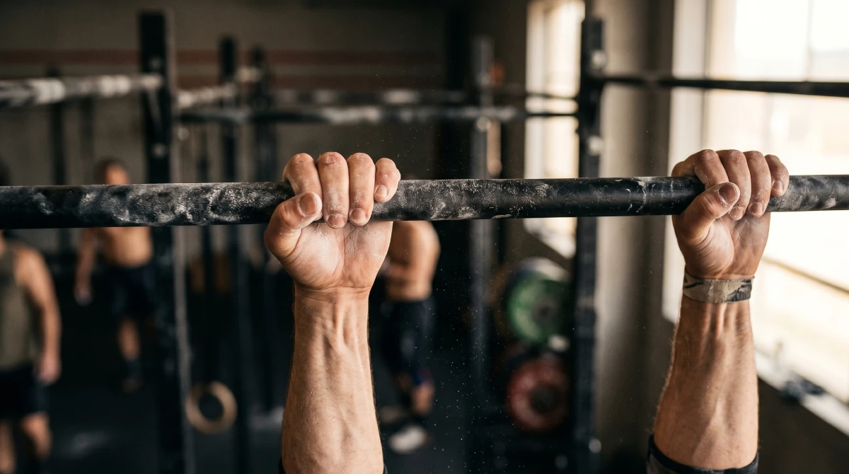 Close-up of athlete's chalked hands gripping a pull-up bar during Marine Corps PFT calisthenics training, illustrating the grip endurance required for high-rep dead-hang pull-up sets